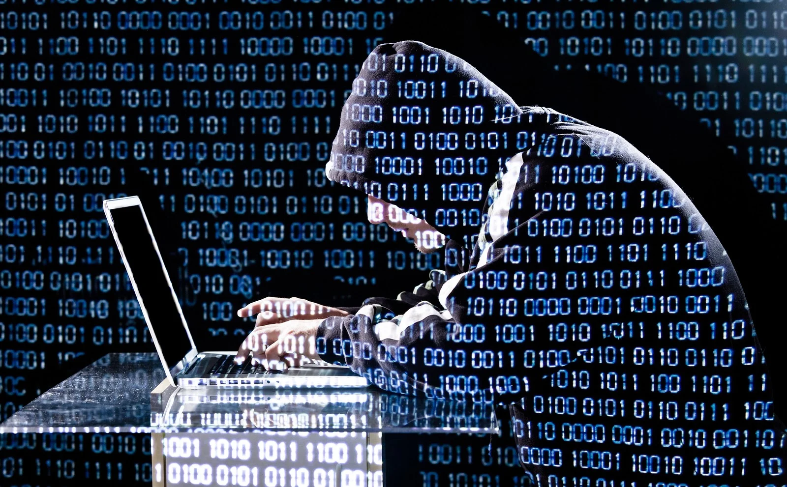 Foto de um homem de capuz usando um laptop. Ele é um esteriótipo de um hacker. Sobre toda foto há a projeção de luzes pintando diversas vezes os digitos 0 e 1.