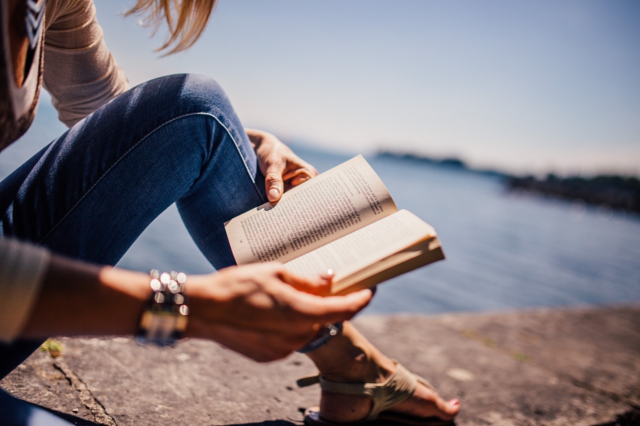foto de uma mulher lendo um livro. Aparece apenas uma de suas pernas e duas mãos. Ela está sentada em um parapeito. Ao fundo podemos ver o oceano