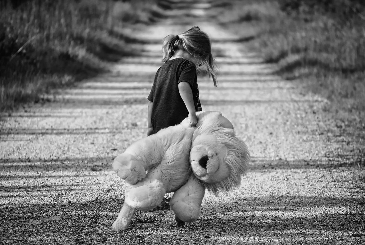 foto preto em branco com uma menina de costas e cabisbaixa segurando um grande urso de pelúcia, arrastando-o no chão. Ela está numa estrada de terra com relva dos dois lados.