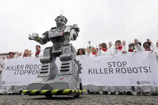 fotos de um protesto. Diversas pessoas ao fundo segurando uma faixa onde se lê: "Stop Killer Robots" (parem os robôs assassinos). A frente dos manifestantes há um robo cinza.
