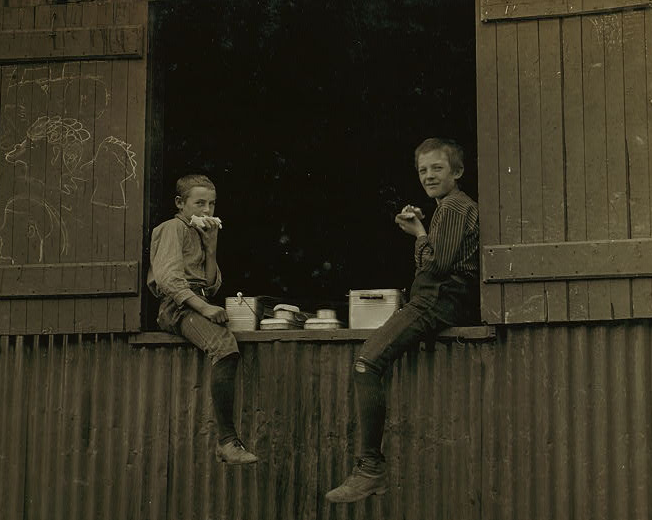 Dois homens comem de suas marmitas, sentados em cima de um vagão de cargas. Pelas roupas usadas, a foto parece ser bastante antiga